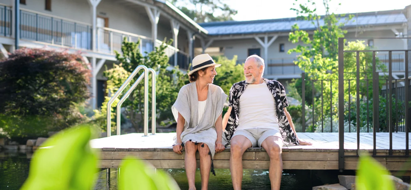 An elderly couple sits on the edge of a wooden deck with their feet in a pond, smiling at each other, in a sunny outdoor setting with buildings and greenery in the background.